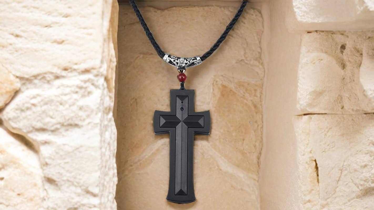 Black cross necklace with red bead hanging on stone wall background.