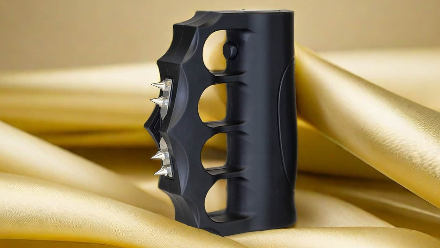 Black self-defense knuckle stun device with spikes on satin background.